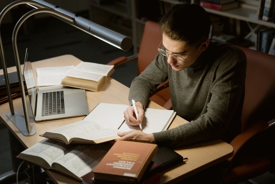 A PDF Workflow for Students: Assignments, Thesis Chapters, and Submissions A young adult studying with books and a laptop, focused on taking notes at a desk under a lamp.