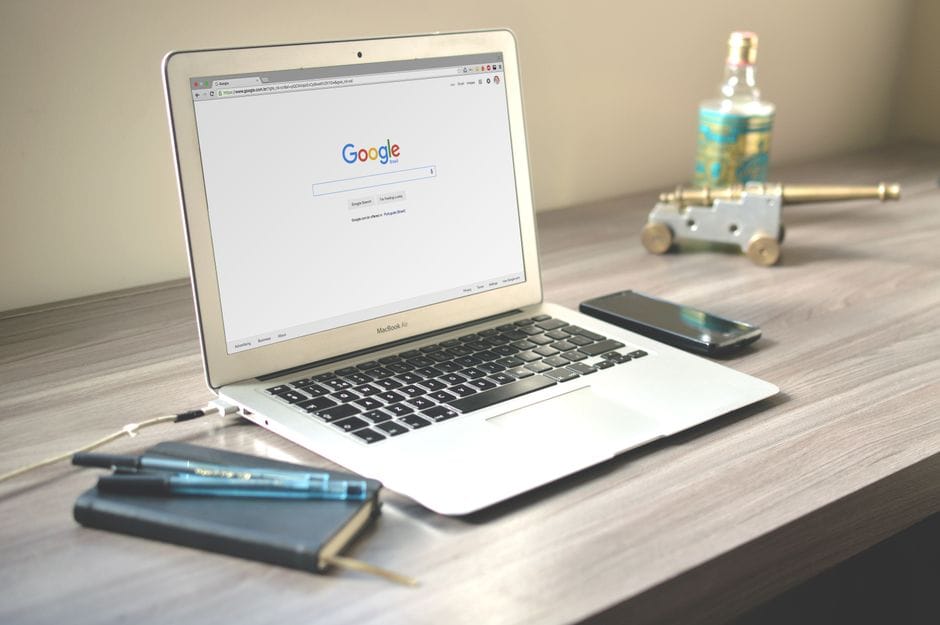 How to Combine PDF Files Without Adobe Acrobat A neat workspace featuring a laptop displaying Google search, a smartphone, and a notebook on a wooden desk.